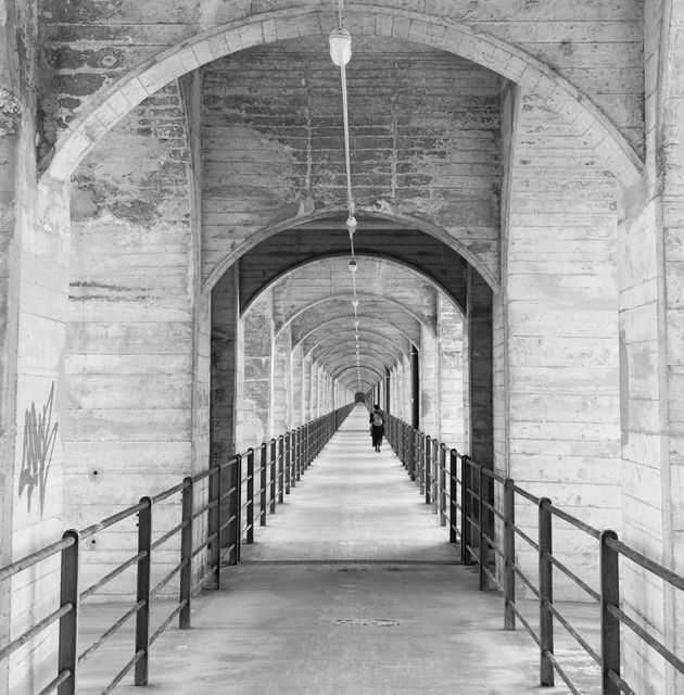 Black and white scene of a solitary figure walking through a long corridor with repeating concrete arches. Captures essence of loneliness and architectural beauty. Perfect for themes of isolation, architecture, urban exploration, or artistic composition.