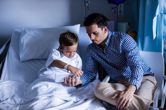 Father and son sitting on hospital bed using digital tablet. Ideal for healthcare, family bonding, pediatric care, and technology in medical settings. Can be used in articles, blogs, and advertisements related to patient care, family support, and hospital experiences.