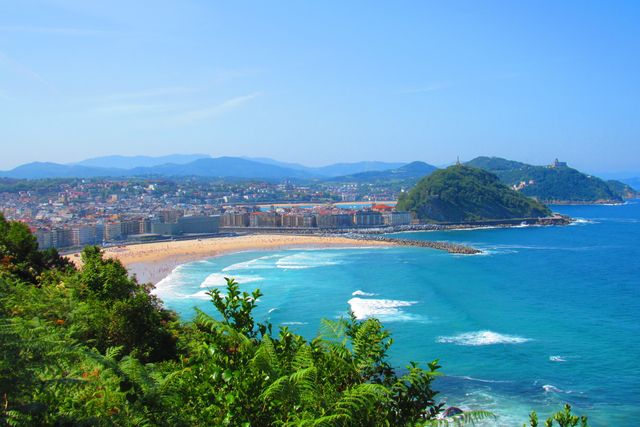 Vibrant image showcasing a coastal city with a beautiful sandy beach and blue ocean waves, framed by green hills and lush foliage under a clear blue sky. Ideal for travel brochures, advertisements, vacation planning, websites, and magazines focusing on tourism and coastal locations.