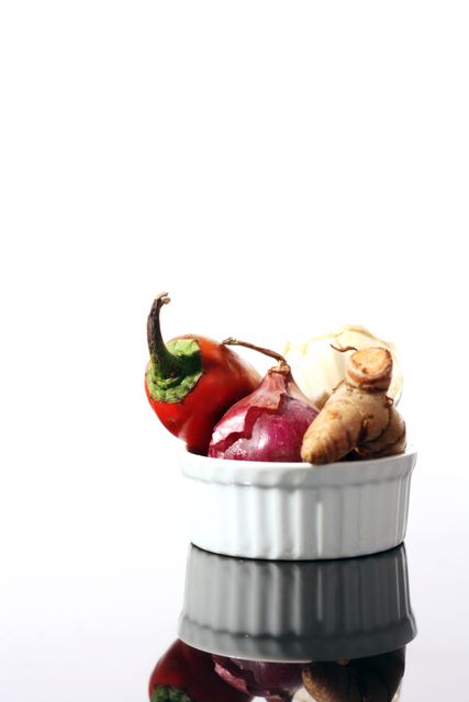 Fresh vegetables and spices including garlic, red onion, red pepper, and ginger root displayed in a white ceramic bowl on a reflective surface. Perfect for use in cooking blogs, recipes, culinary websites, and food-related advertisements showcasing fresh and healthy ingredients.