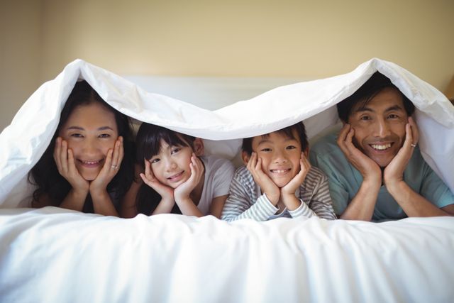 This image shows a happy family relaxing together under a blanket in a bedroom at home. The parents and children are smiling and bonding, creating a cozy and loving atmosphere. This image can be used for promoting family values, home comfort, and togetherness. It is ideal for advertisements, family-oriented blogs, and social media posts highlighting family life and happiness.