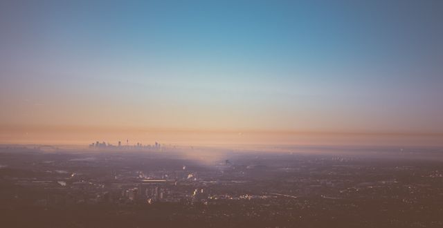 Panoramic view of a cityscape during sunrise with a clear sky and colorful hues. Buildings create a dotted pattern over the horizon, and light mist adds a serene atmosphere. Ideal for use in travel blogs, urban landscape posters, environmental articles emphasizing air quality, or any content related to city life and morning ambiance.