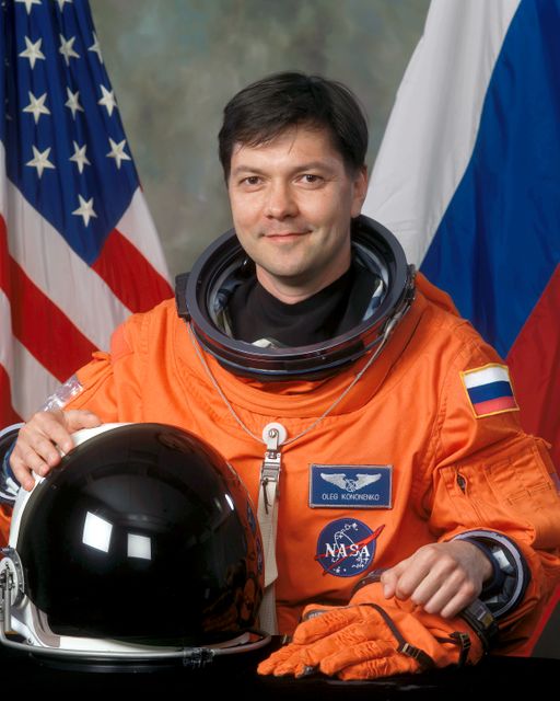 This image shows cosmonaut Oleg Kononenko from Russia's Federal Space Agency wearing an orange space suit and holding a space helmet. The Russian and American flags are in the background, emphasizing international collaboration in space exploration. This photograph is ideal for use in articles about space missions, astronaut training, and international space cooperation. It can also be used for educational purposes related to space studies.