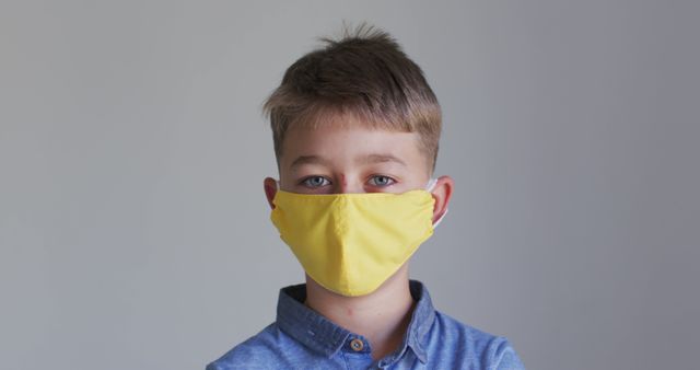 Young boy wearing yellow face mask and blue shirt standing against a neutral background. Represents health and safety measures during a pandemic. Suitable for topics related to child healthcare, pandemic precautions, Covid-19 awareness, school safety, and public health campaigns.
