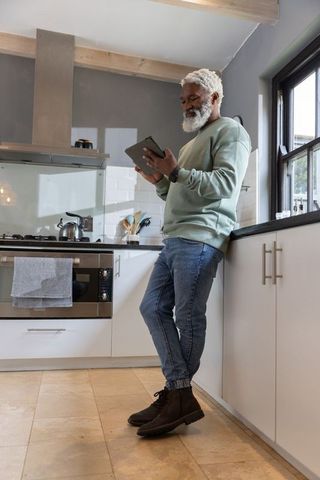 Senior man leaning in modern kitchen using tablet, casual sweatshirt and wristwatch