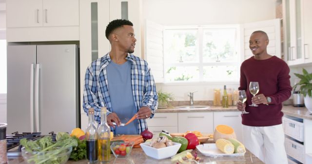 Two African American men are preparing a healthy meal together in a bright, modern kitchen. One is cutting vegetables while the other holds a glass of white wine. This image can be used to illustrate themes of healthy living, friendship, domestic life, or home cooking. It is suitable for advertisements, blog posts about healthy eating, or articles on cooking and lifestyle.