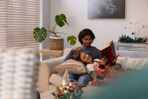 Diverse couple reclining on sofa reading red hardcover book together during cozy holiday evening