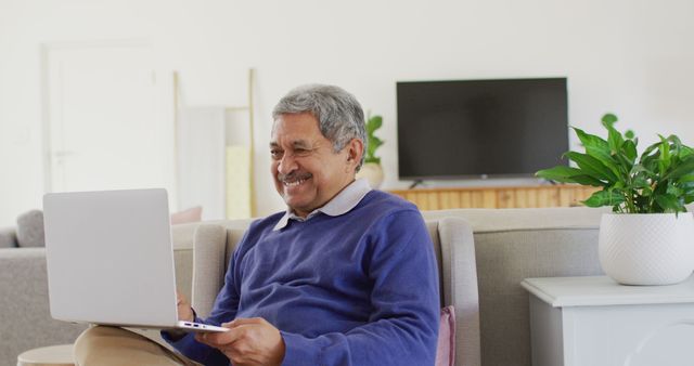 An elderly man sits comfortably on a couch while using a laptop in a bright and modern living room. He is smiling, indicating happiness and satisfaction. This image is ideal for use in articles or advertisements related to technology for seniors, remote communication, or promoting a relaxed and comfortable lifestyle for the elderly.