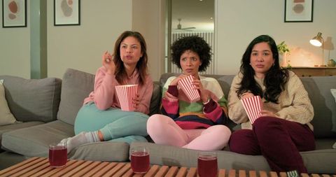 Multicultural women watching movie on sofa, eating popcorn and enjoying cozy night in