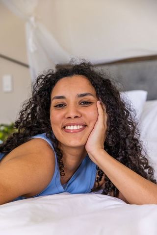 Smiling african american woman reclining on bed wearing blue top in bright minimalist bedroom