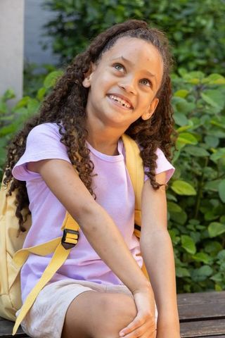 Joyful schoolgirl smiling while sitting on bench wearing yellow backpack in sunlit garden