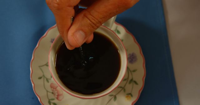 A hand stirring coffee in a floral teacup placed on a blue table setting. Ideal for use in ads or articles about morning routines, coffee culture, relaxation time, or editorial pieces focusing on simple daily rituals.