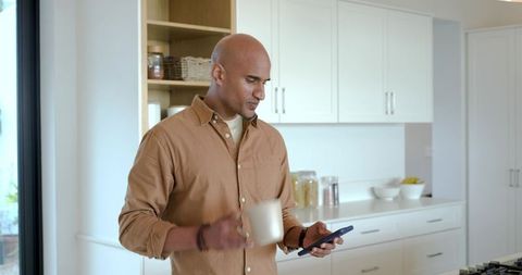 Indian man holding coffee mug and checking smartphone in bright minimalist kitchen