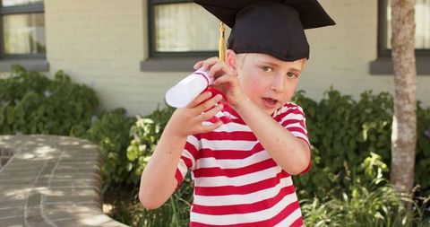 Young boy celebrating graduation wearing mortarboard and holding diploma outdoors