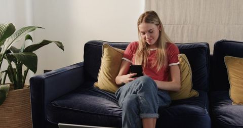 Young woman relaxing on navy sofa using smartphone and laptop in cozy living room