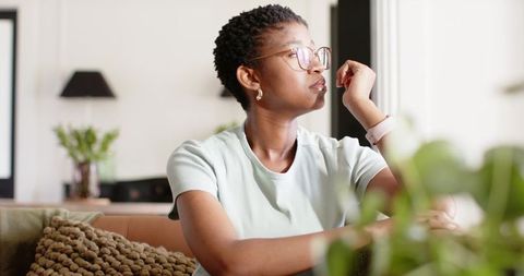 Woman gazing out window on sunlit sofa with houseplants and cozy modern living room