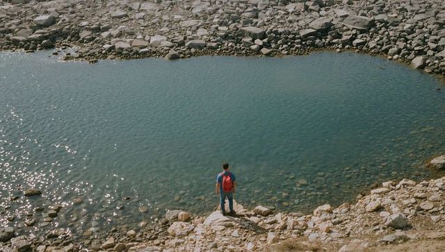 Standing hiker overlooking clear mountain pond on rocky lakeshore with red backpack