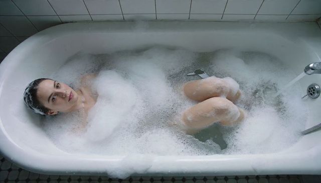 Woman relaxing in bubble bath in vintage enamel bathtub with foam and serene expression, home spa