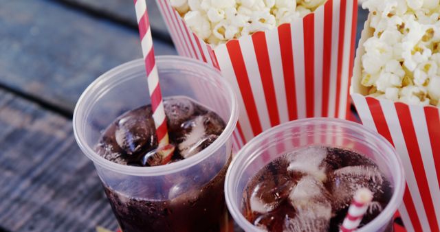 Shows two cups of cold drinks with ice and striped paper straws next to striped cartons of popcorn on a wooden table. Ideal for illustrating summer picnics, movie nights, parties, and snack time themes.