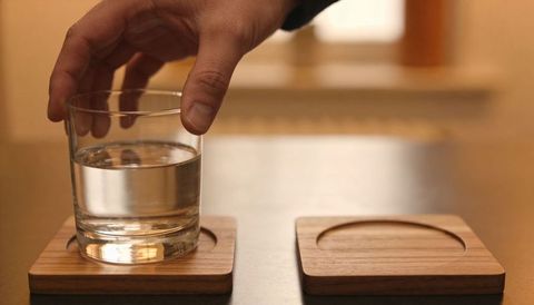 Placing clear glass of water on square wooden coaster on warm tabletop closeup