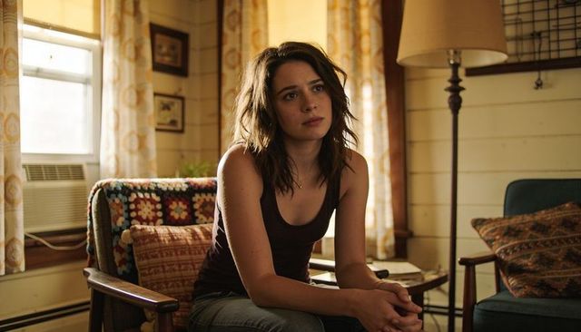 Pensive young woman sitting in sunlit living room wearing dark tank top and jeans