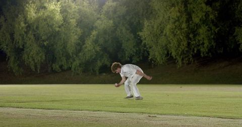 Cricket fielder bending to pick up ball on night outfield under stadium lights