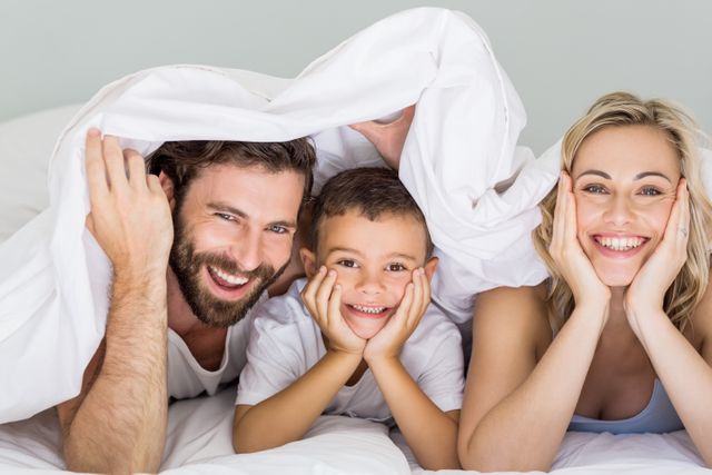 This image shows a happy family consisting of parents and their son lying on a bed under a blanket, all smiling warmly. It is perfect for use in advertisements, family-oriented blogs, parenting articles, and lifestyle magazines to depict family bonding, joy, and relaxation at home.