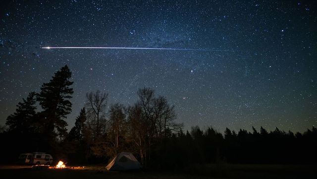 Campfire illuminating tent under starry sky with meteor streak and camper van nightscape