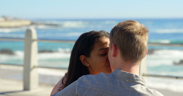 A young biracial couple shares a kiss by the seaside, with copy space. Their affectionate moment captures a romantic and serene beach setting.