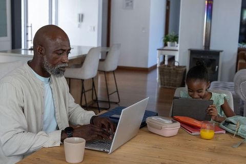 Middle-aged african american father working remotely at table while daughter using tablet