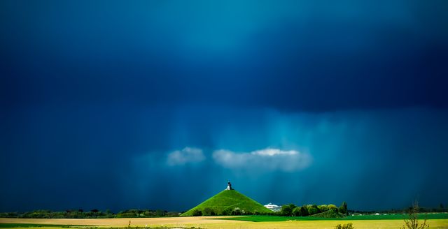 Image showcases a dramatic sky over a lush green landscape with a mound topped with a monument. Suitable for use in travel blogs, nature photography websites, and digital art backgrounds depicting stormy weather scenes.