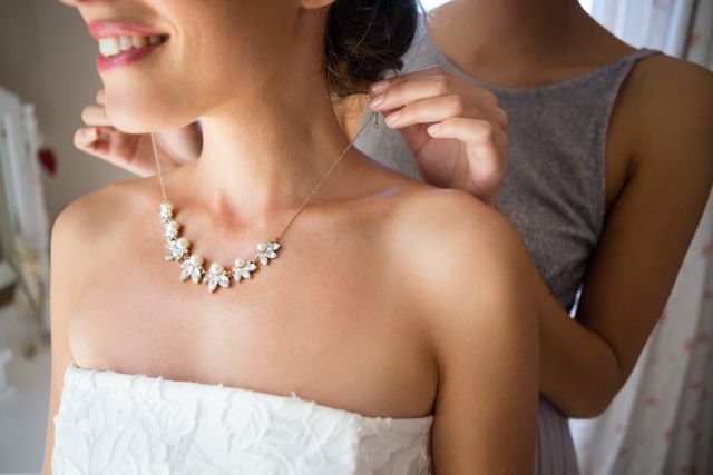 Bridesmaid fastening a beautiful necklace around the bride's neck in a dressing room. Ideal for use in wedding planning materials, bridal magazines, jewelry advertisements, and articles about wedding preparations and ceremonies.