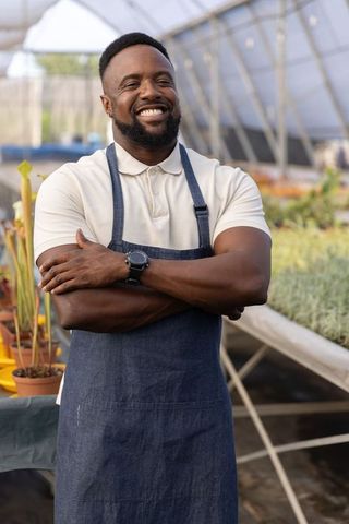 African american nursery owner smiling in greenhouse wearing denim apron and watch