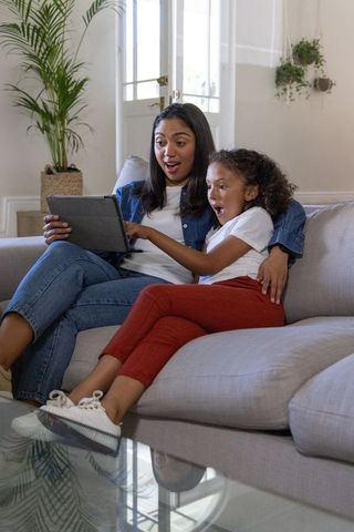 Mother and daughter sharing tablet on light-gray sofa, reacting with surprise at home