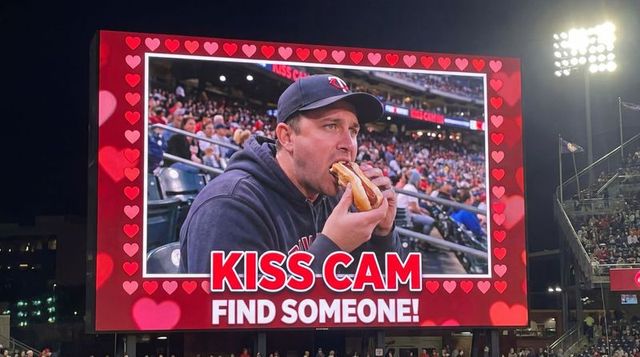 Kiss cam moment man eating hot dog on jumbotron at night baseball game crowd cheering