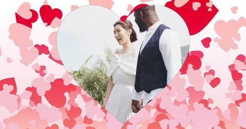 Multicultural couple laughing and holding hands in meadow through heart cutout with pink hearts