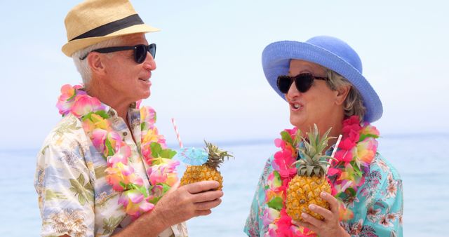 Elderly couple enjoying a tropical vacation on a sunny beach, wearing leis and straw hats, smiling and holding pineapples decorated with drink umbrellas. Ideal for concepts related to travel, retirement, vacations, enjoyment, and healthy lifestyles for seniors.