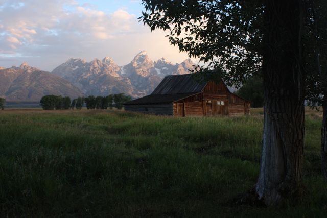 Rustic barn sitting in grassy field with a backdrop of majestic mountain ranges at sunset creating a serene and peaceful atmosphere. Suitable for illustrating rural life, nature retreats, travel blogs, picturesque landscapes, and promotional materials for outdoor activities and adventures.