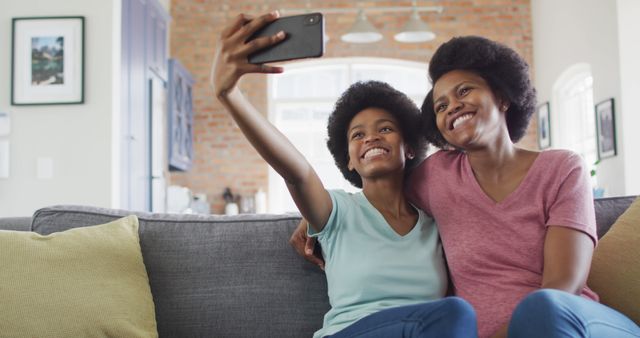 Happy african american mother and daughter sitting on sofa using smartphone, taking selfie. domestic life and quality family time together at home.
