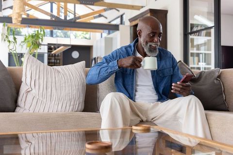 Senior african american man sipping coffee and scrolling phone on sofa in modern living room