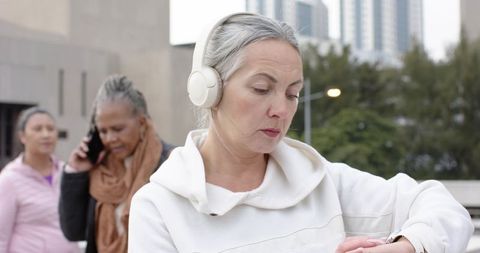 Mature woman wearing headphones checking smartwatch while walking in urban plaza