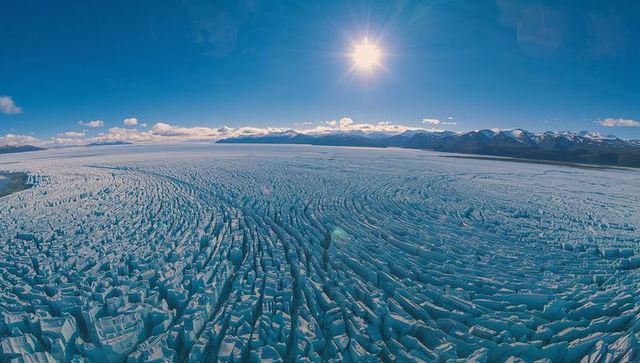 Fracturing glacier radiating seracs and crevasses toward distant mountains under sun