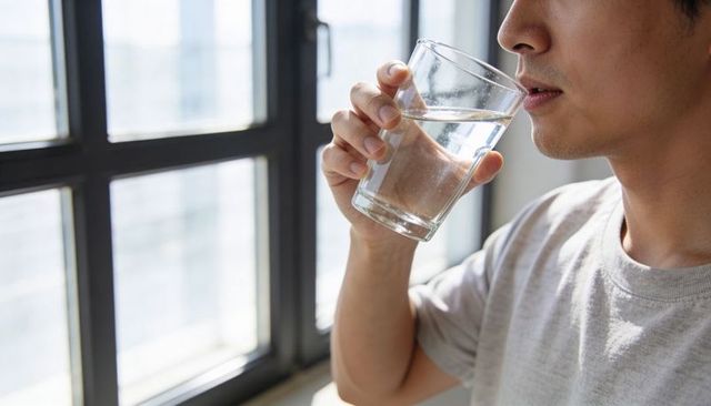 Asian man drinking water by metal-framed window, holding glass tumbler, serene daylight