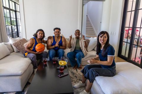 Friends watching basketball game at home cheering on sectional couch holding basketball, snacks, dri
