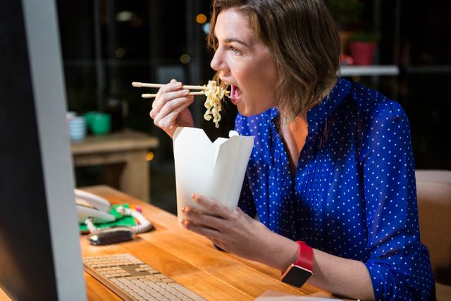 Businesswoman eating noodles at her desk in the office, using chopsticks. Ideal for illustrating concepts of busy work life, late-night work, multitasking, and modern office environments. Useful for articles or advertisements related to work-life balance, office culture, and productivity.