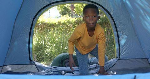 African american boy crawling out of blue tent in sunny backyard during family camping trip