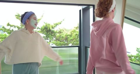 Diverse friends enjoying skincare ritual on sunny balcony wearing hoodies and face masks
