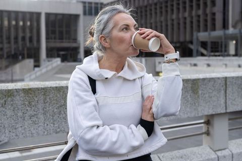 Mature woman drinking takeaway coffee in city plaza wearing white hoodie and smartwatch