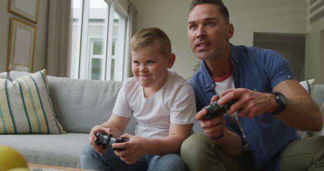 Father and son are sitting on the sofa in the living room playing video games together. Both are holding game controllers and focused on the game; the boy looks particularly excited. The scene is ideal for illustrating family bonding, leisure activities, father's day content, or technology use in home settings.