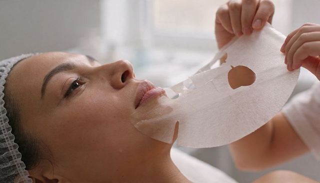 Esthetician applying hydrating sheet mask on female client during relaxing facial treatment